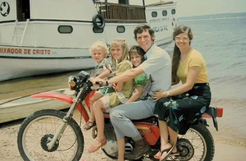 Lucas, Christine e os três filhos numa moto à beira do rio, com um barco da missão ao fundo