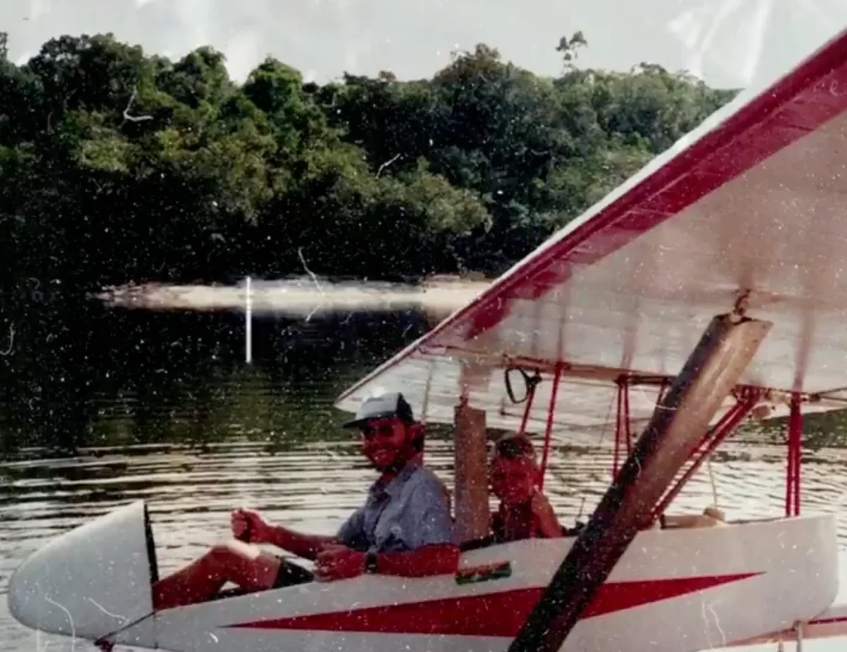 O Pastor Lucas Huber sorrindo no pequeno avião branco e vermelho, pousado no rio, com a mata ao fundo